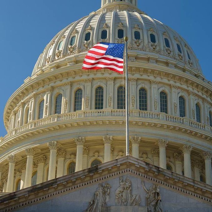 The dome of the United States Capitol building with an American flag flying in front of a blue sky.