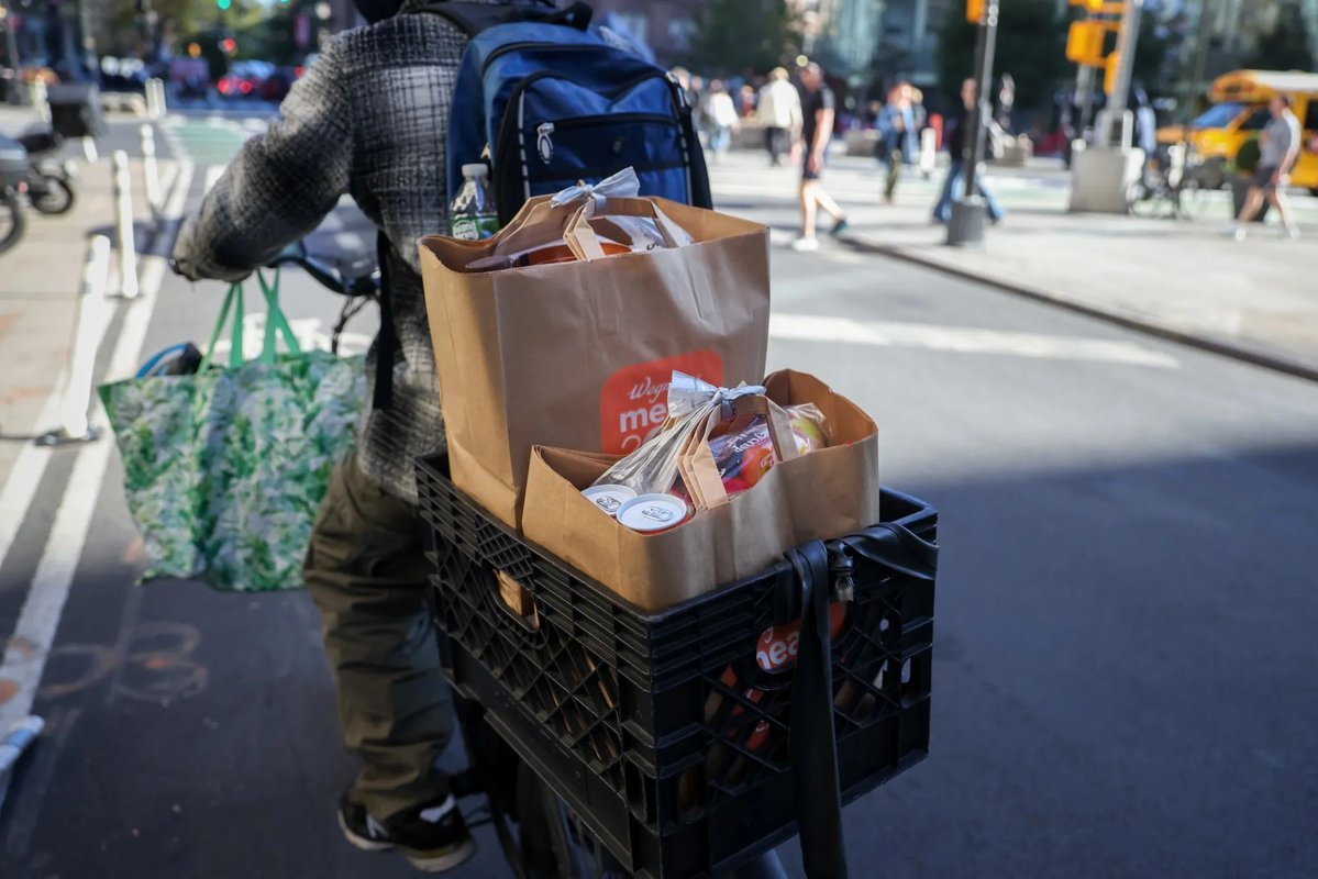 A delivery courier on a bicycle carrying paper grocery bags in a rear crate on a city street.