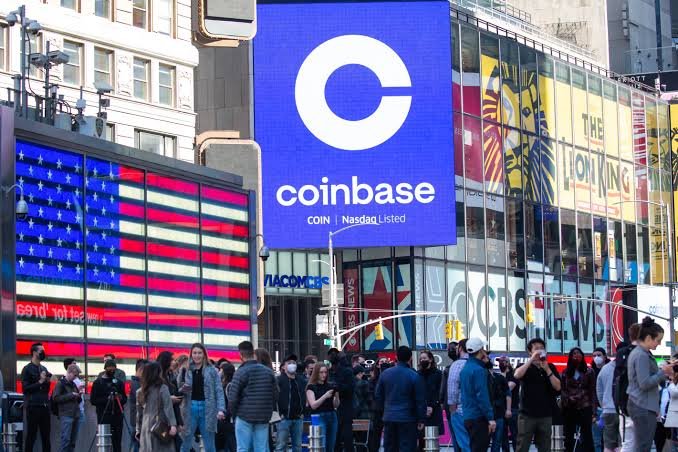 Pedestrians walk past a large outdoor blue digital billboard displaying the Coinbase logo and name in a city square.