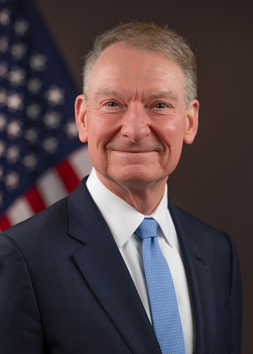 Portrait of SEC Chair Atkins smiling in a suit and tie in front of an American flag.