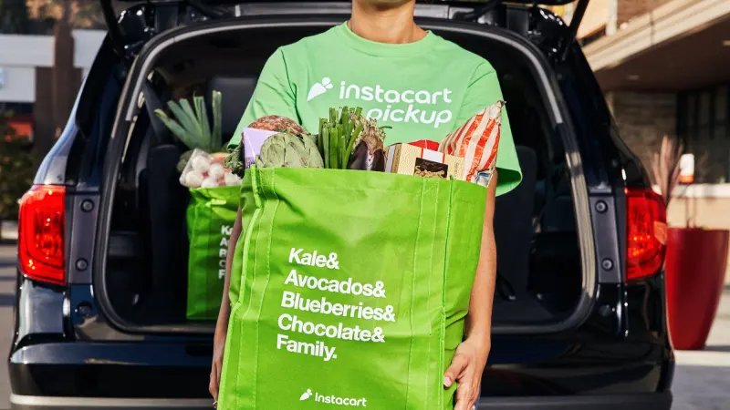 An Instacart delivery worker holding a reusable green grocery bag loaded with fresh produce and food items.