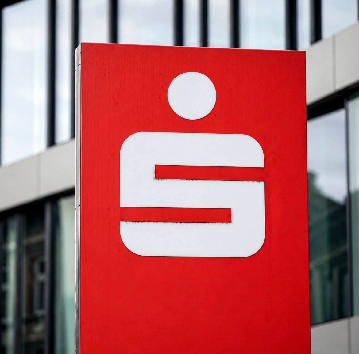 A red Sparkassen sign with its white 'S' logo displayed in front of a modern building.