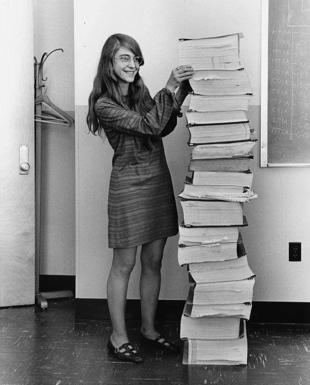 Margaret Hamilton smiling as she stands next to a tall vertical stack of bound Apollo navigation software code listings.