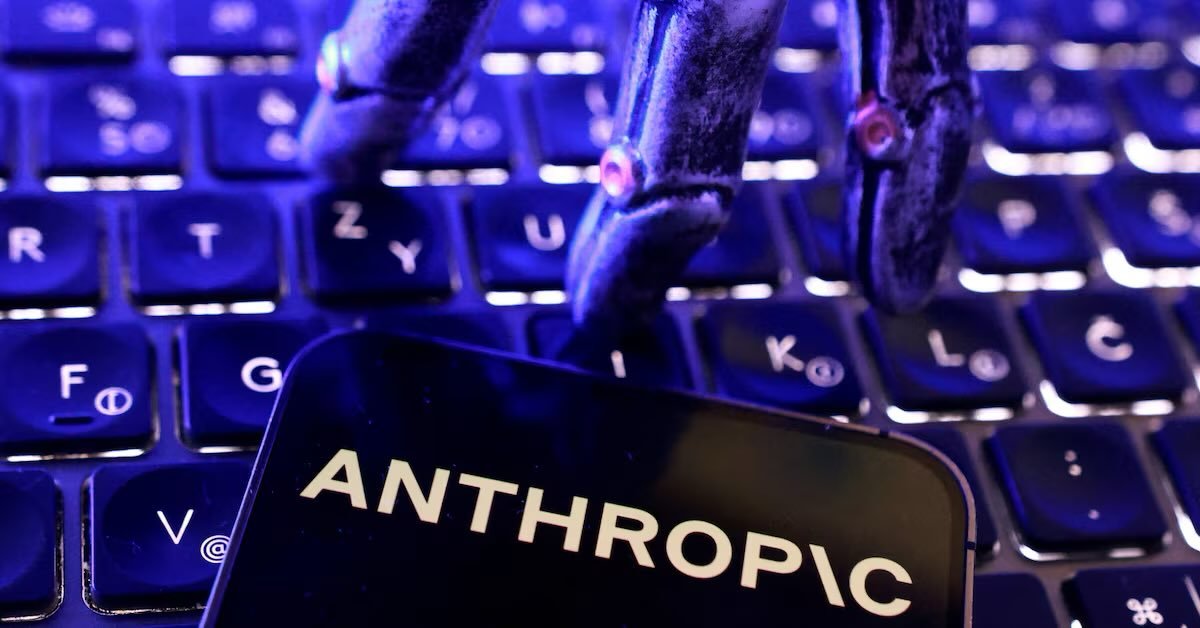 A robotic hand types on a backlit keyboard next to a smartphone showing the Anthropic logo.