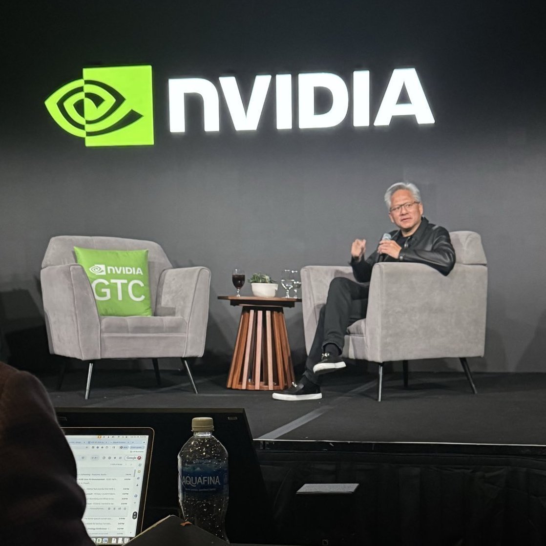 Nvidia CEO Jensen Huang speaking on stage in an armchair beneath an illuminated Nvidia logo.