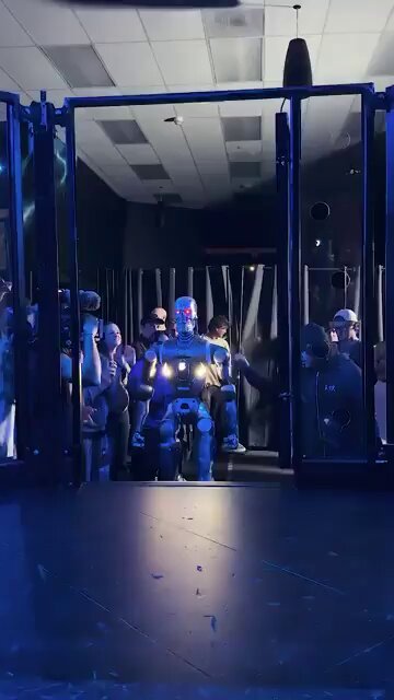 A metallic humanoid robot with glowing red eyes, styled like a Terminator, enters through a doorway into an arena surrounded by spectators.