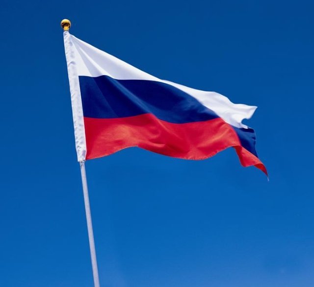 The Russian national flag waving on a flagpole against a clear blue sky.