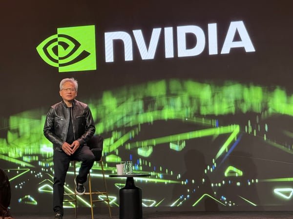 Nvidia CEO Jensen Huang sits on a stool on stage in front of a large screen displaying the Nvidia logo.