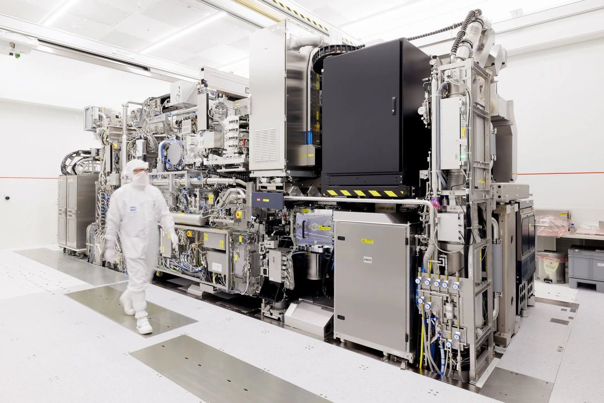 A technician in cleanroom gear walks alongside a large ASML semiconductor lithography machine in a fabrication facility.