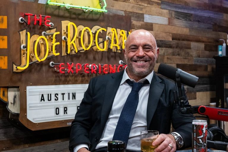 Joe Rogan smiling in a suit and tie while seated in front of a microphone and The Joe Rogan Experience neon sign in his studio.