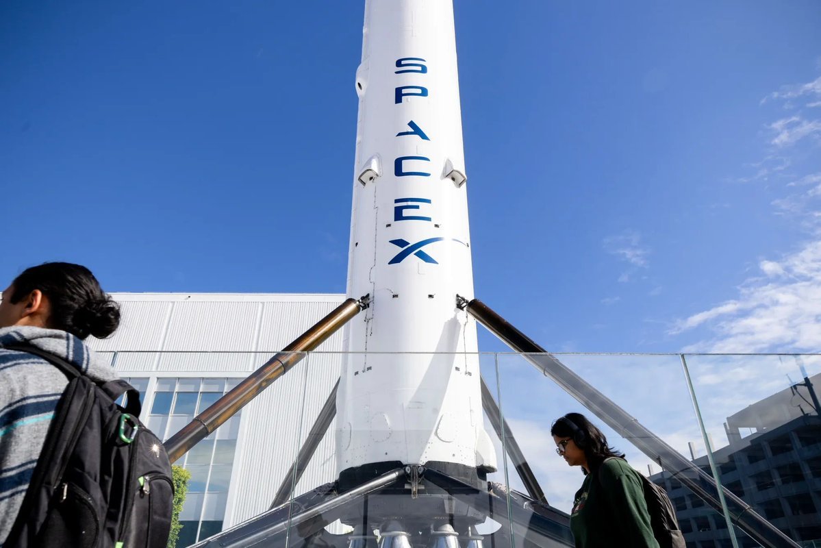 A SpaceX Falcon 9 rocket booster on display outside a company facility with pedestrians walking past.