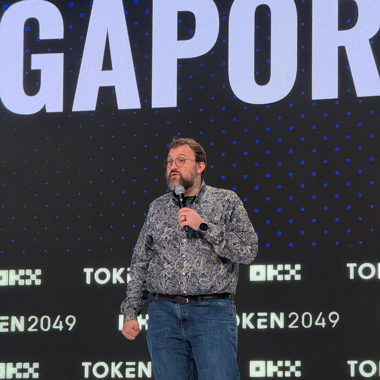 Charles Hoskinson speaks into a microphone on stage in front of a screen displaying TOKEN2049.