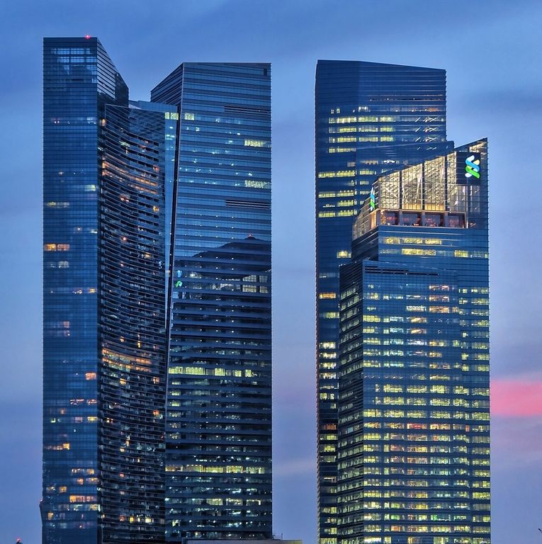 Modern illuminated skyscrapers at dusk, including the Marina Bay Financial Centre bearing the Standard Chartered logo in Singapore.