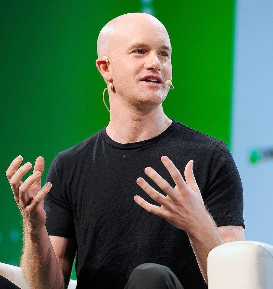 Coinbase CEO Brian Armstrong speaking on stage in a black T-shirt against a green background.