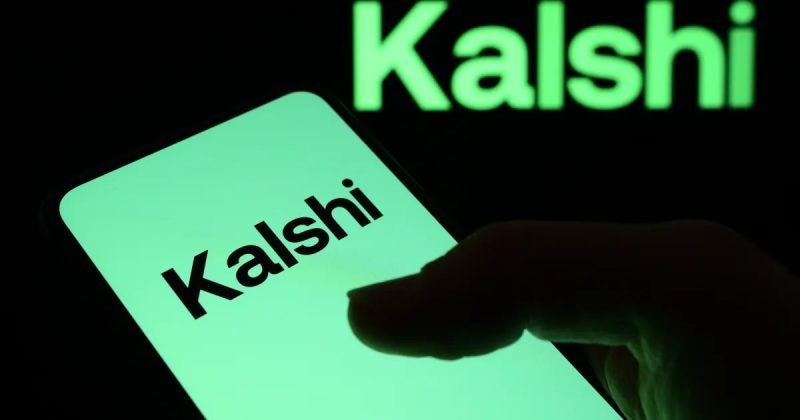 A smartphone displaying the Kalshi logo in front of a monitor showing the same logo.