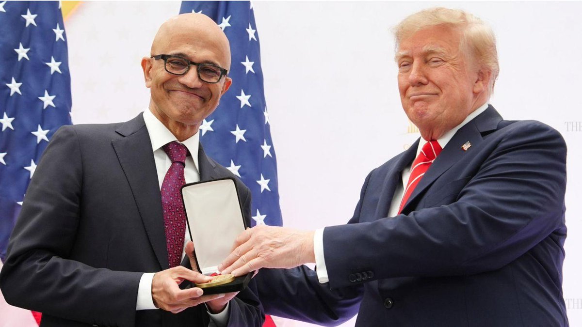 Donald Trump presenting an award medal to Microsoft CEO Satya Nadella in front of American flags.