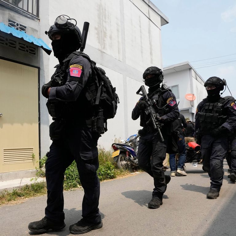 Tactical police officers wearing black uniforms, helmets, and face coverings walk in gear along a street outside buildings.