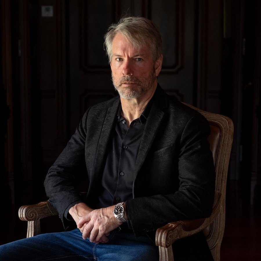 Portrait of Michael Saylor sitting in a wooden chair wearing a black shirt and jacket.