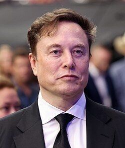 Portrait of Elon Musk wearing a dark suit and tie.