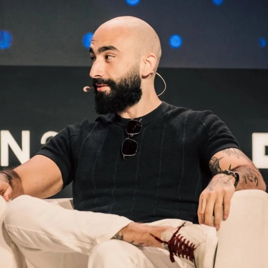 Helius CEO Mert Mumtaz seated on a stage panel wearing a black shirt and white pants.