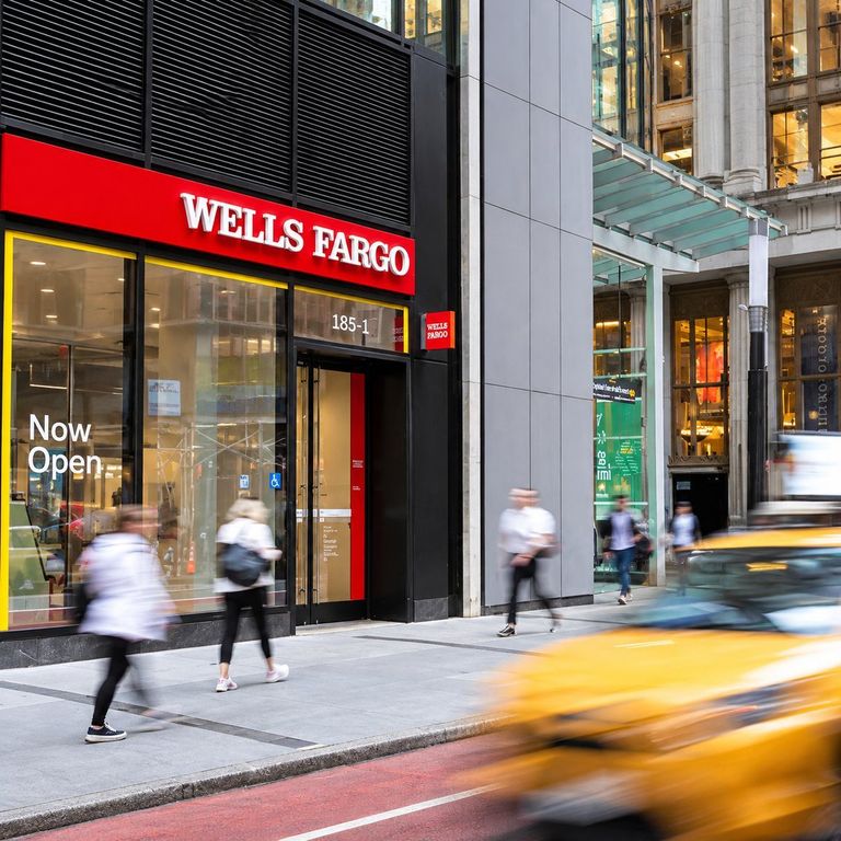 Exterior view of a Wells Fargo branch entrance on a city sidewalk with pedestrians walking by and a yellow taxi driving past in motion blur.