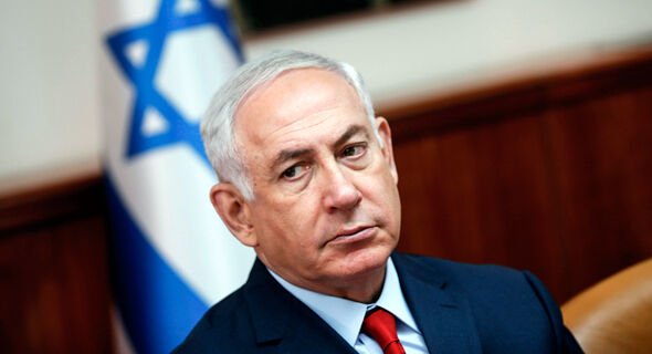 Close-up portrait of Israeli Prime Minister Benjamin Netanyahu seated in front of an Israeli flag.
