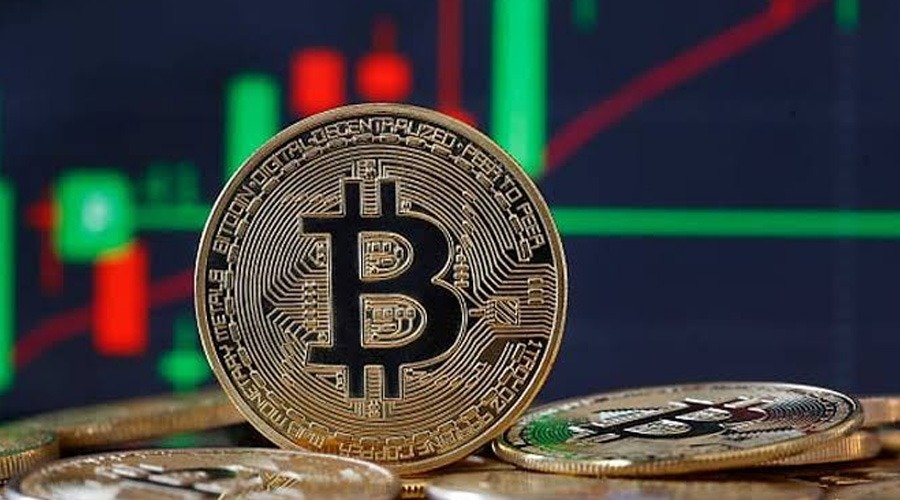 A physical Bitcoin coin resting upright against an out-of-focus background showing red and green financial candlestick charts.