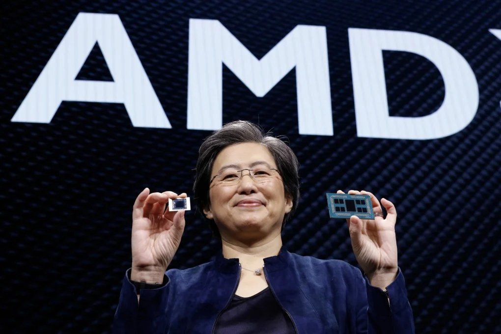 AMD CEO Lisa Su smiling on stage while holding semiconductor chip hardware in front of an AMD logo.