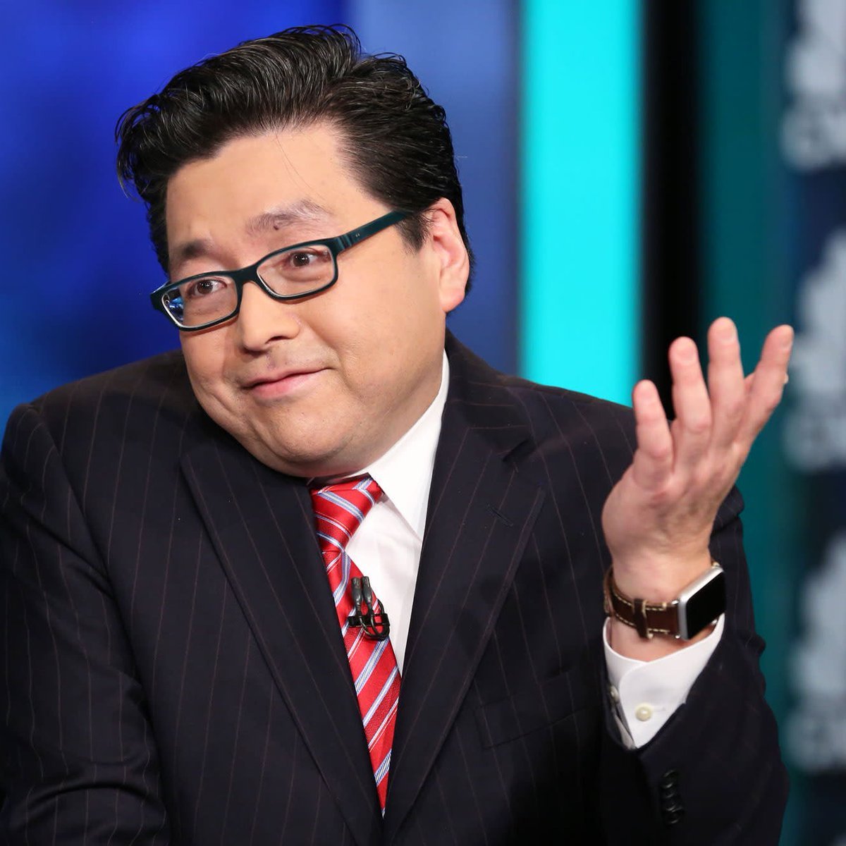 Portrait of Fundstrat co-founder Tom Lee wearing glasses, a suit, and a red striped tie while gesturing with his hand.