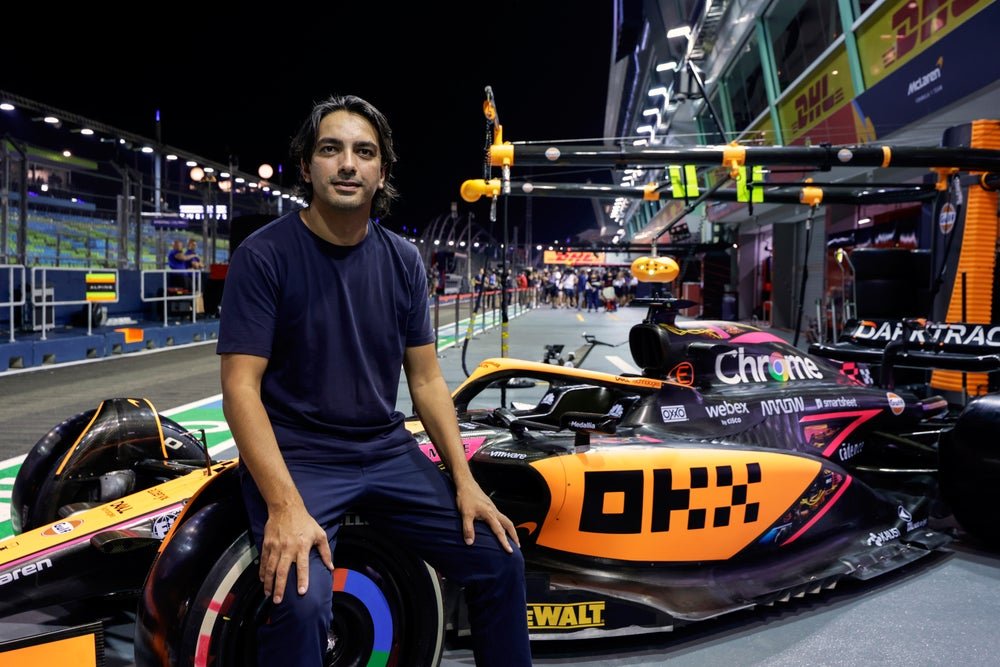 Haider Rafique seated on the tire of an OKX-branded McLaren Formula 1 race car in a pit lane.