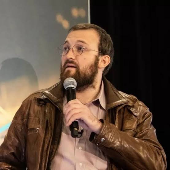 Cardano founder Charles Hoskinson speaking into a microphone while wearing a brown leather jacket and glasses.