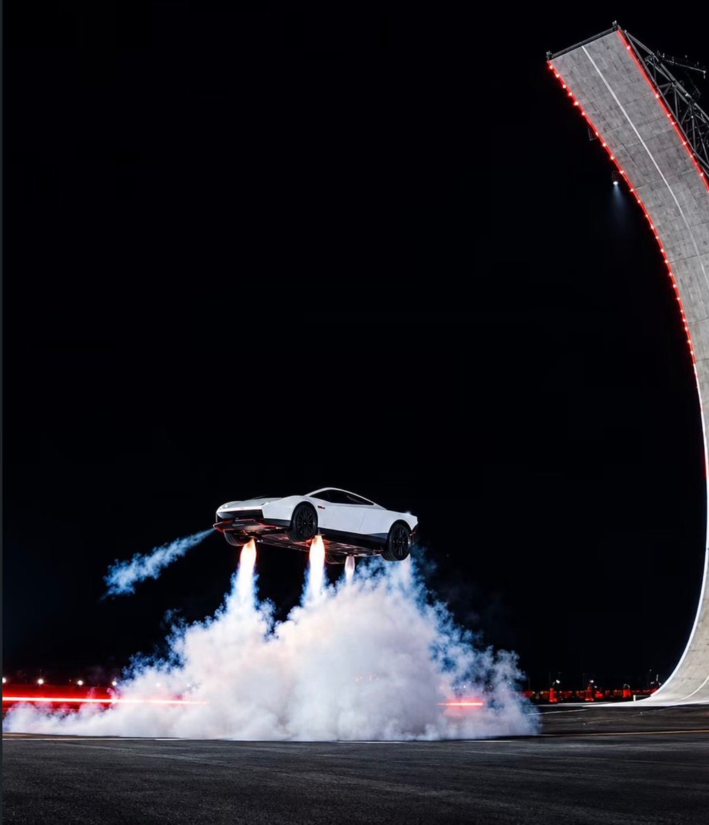 A white Tesla Roadster prototype hovers midair propelled by thrusters emitting smoke next to a tall curved ramp at night.