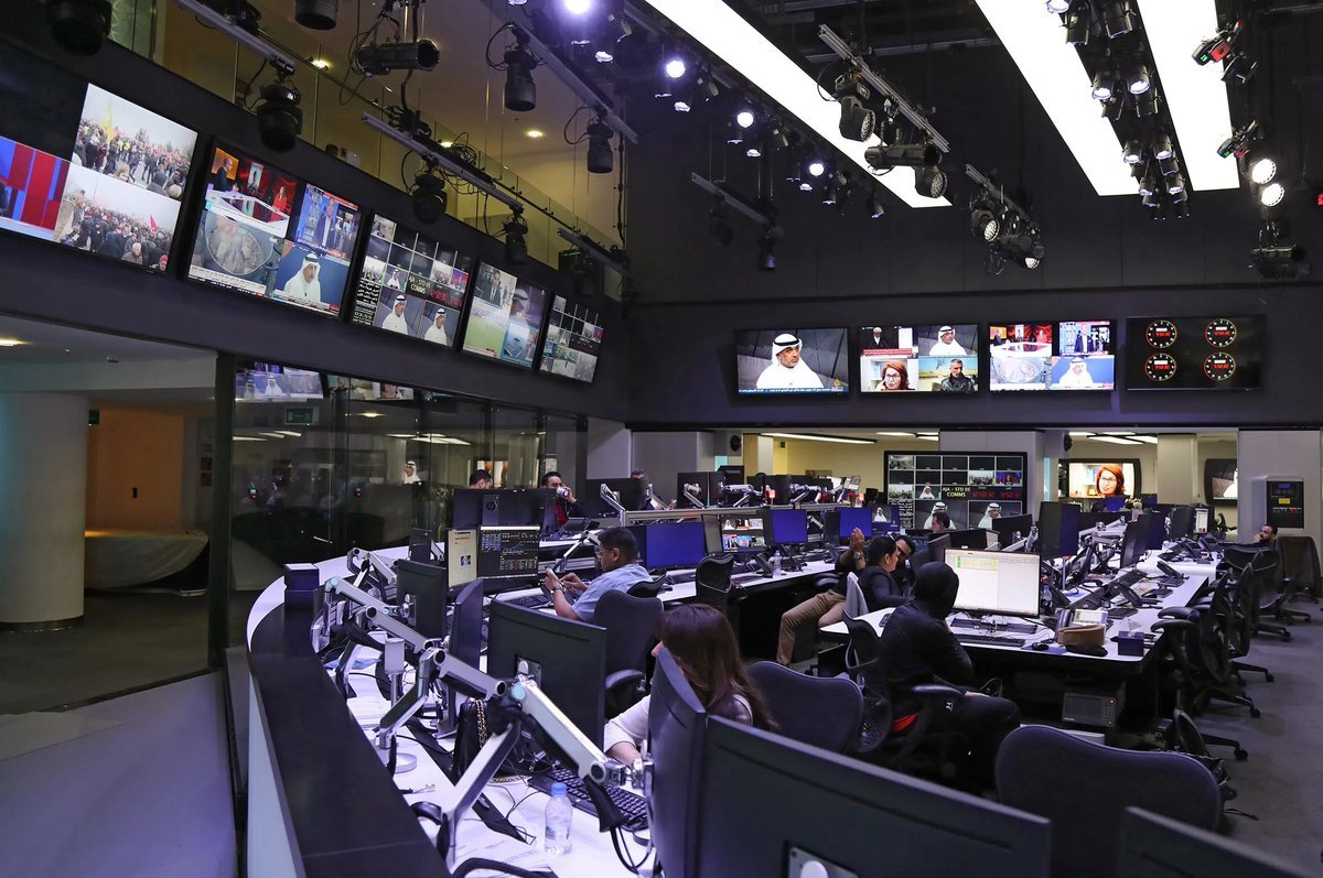 Journalists and staff working at computer terminals inside a newsroom and broadcast control center with television monitors mounted on the wall.
