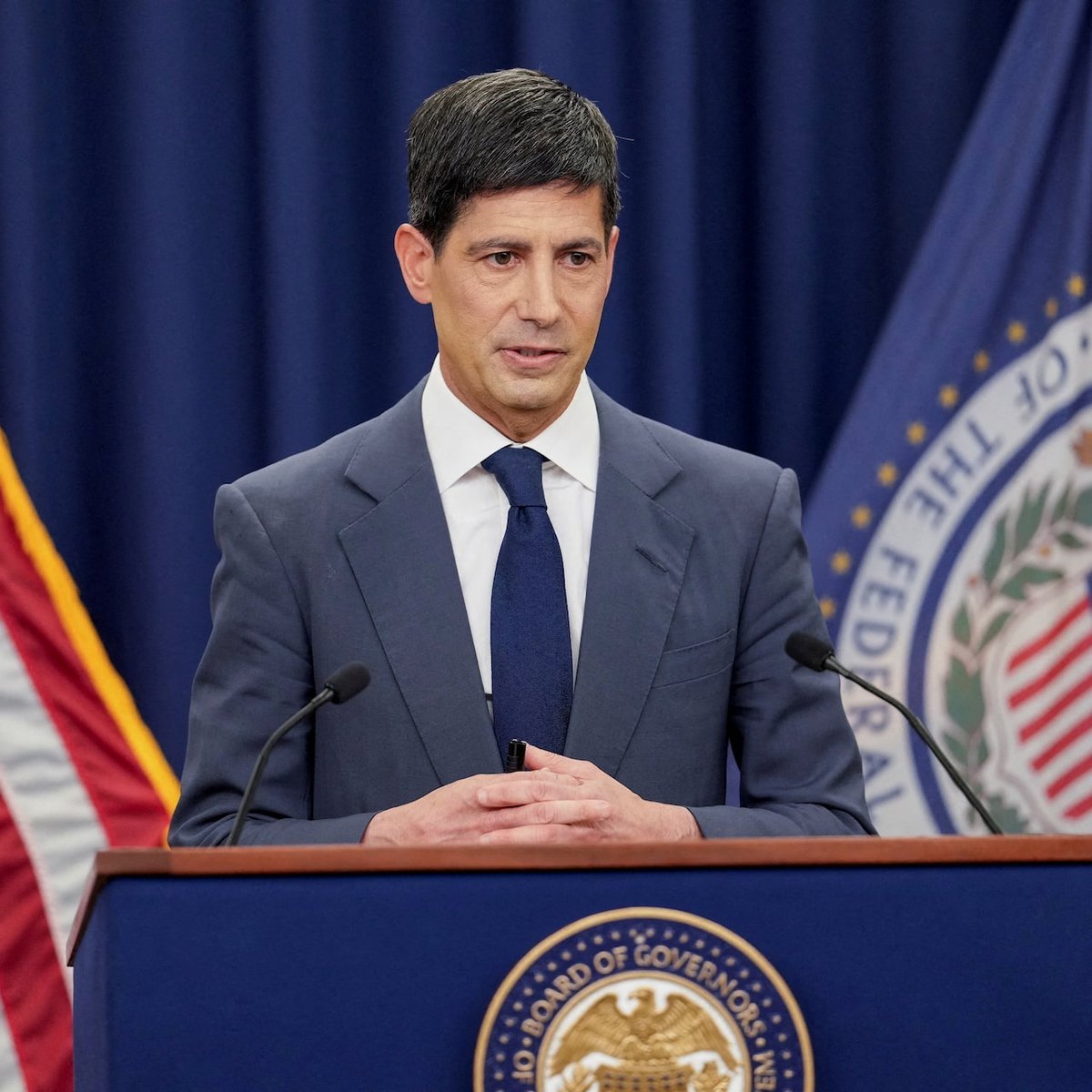 A man in a suit speaking at a podium bearing the seal of the Board of Governors of the Federal Reserve System.