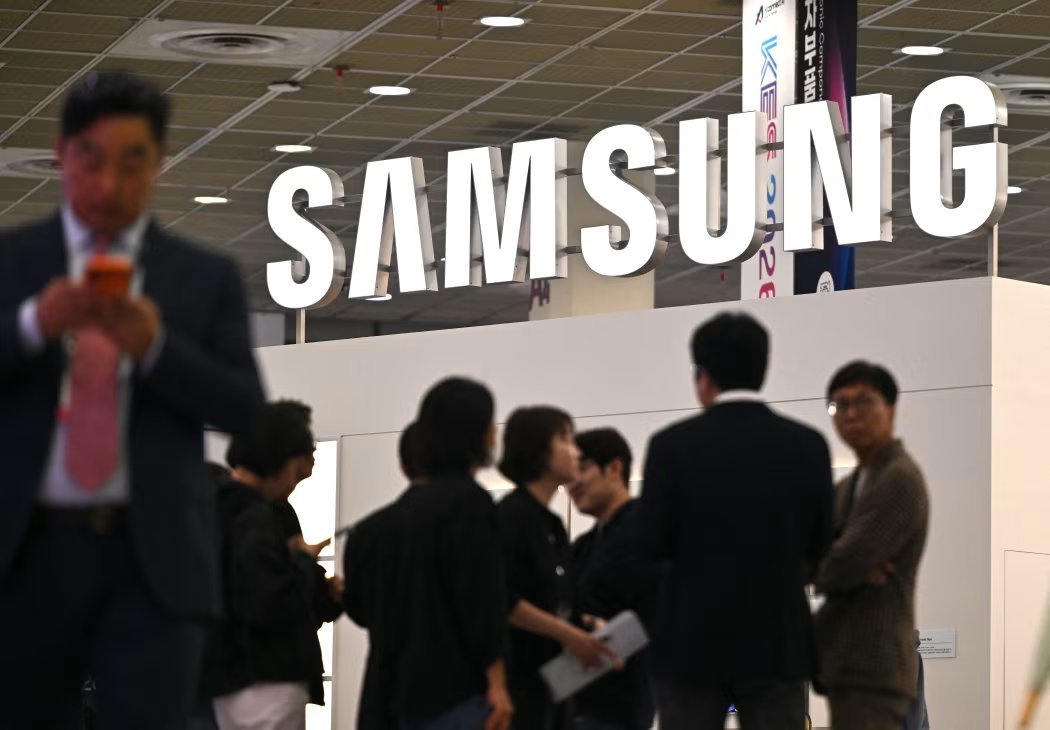 A large illuminated Samsung logo above an exhibition booth with people walking in the foreground.