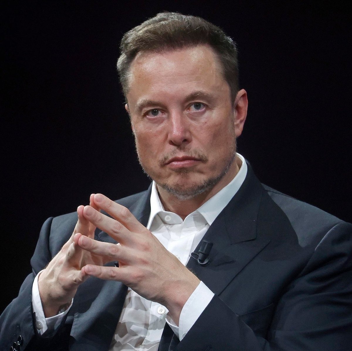 Portrait of Elon Musk wearing a dark suit jacket and white collared shirt.