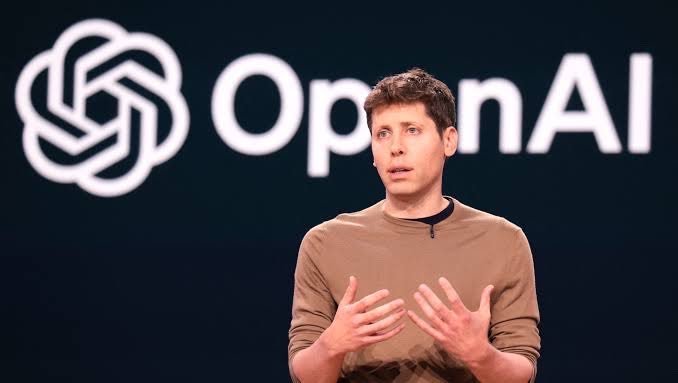 OpenAI CEO Sam Altman gesturing while speaking on stage in front of a large OpenAI logo.