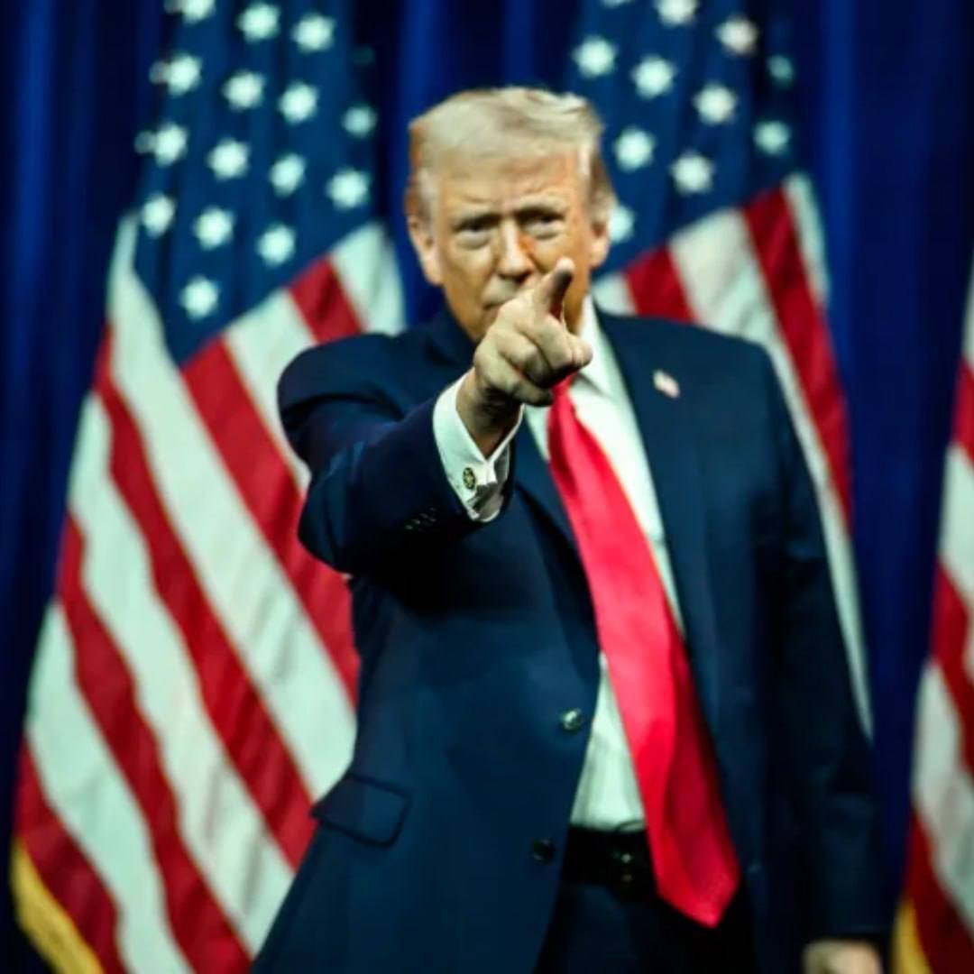 Donald Trump in a dark suit and red tie pointing forward in front of American flags