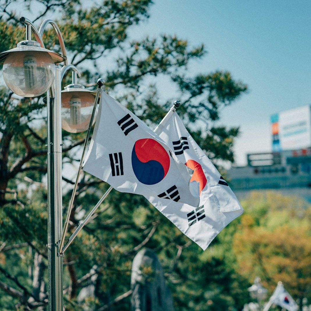 South Korean flags waving on a pole outdoors against trees and a building.