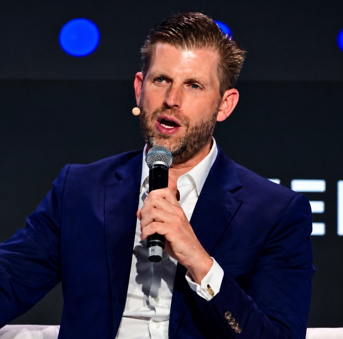 Eric Trump speaks into a microphone while wearing a blue suit jacket and white shirt on a stage.