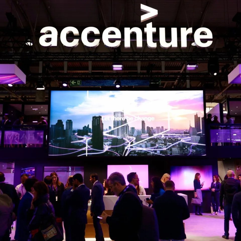 An Accenture exhibition booth with the company logo prominently displayed above a large video screen and attendees below.