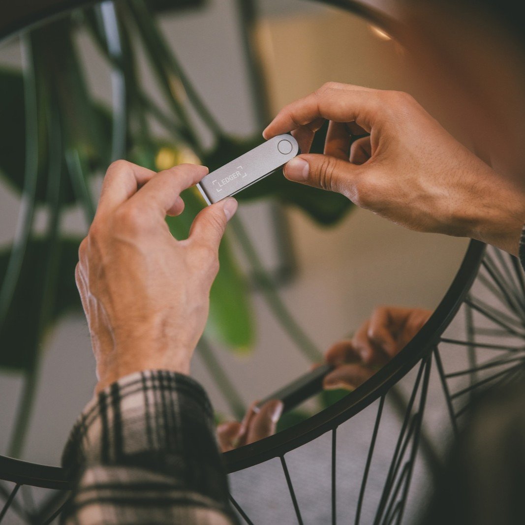 A person holding a small silver Ledger hardware cryptocurrency wallet device.