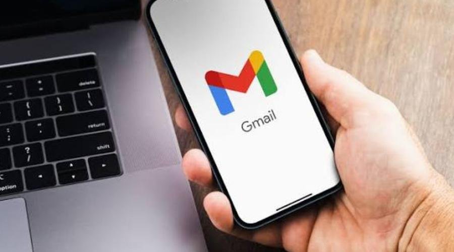 A person holding a smartphone showing the Gmail logo on its screen next to an open laptop.