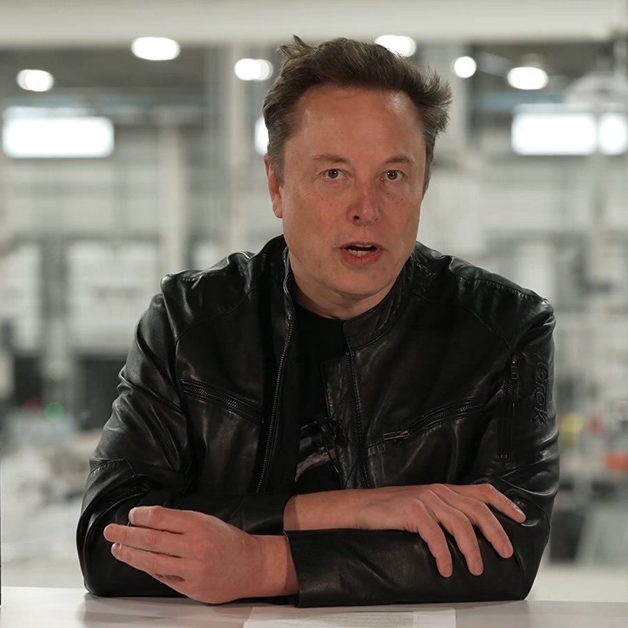 Elon Musk wearing a black leather jacket seated at a table during a broadcast.