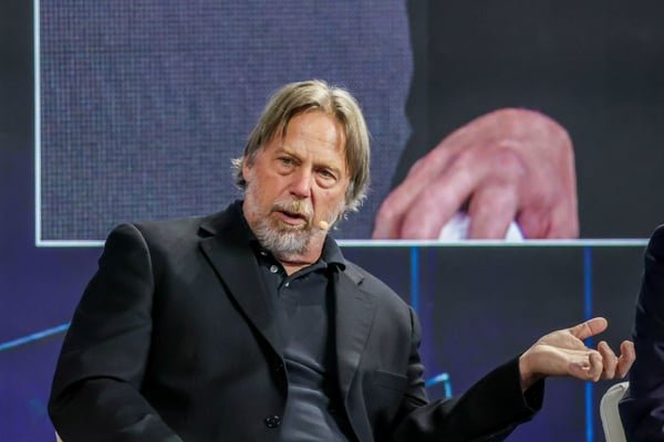 A man with a beard in a black suit jacket speaks while gesturing with his hand on a stage.