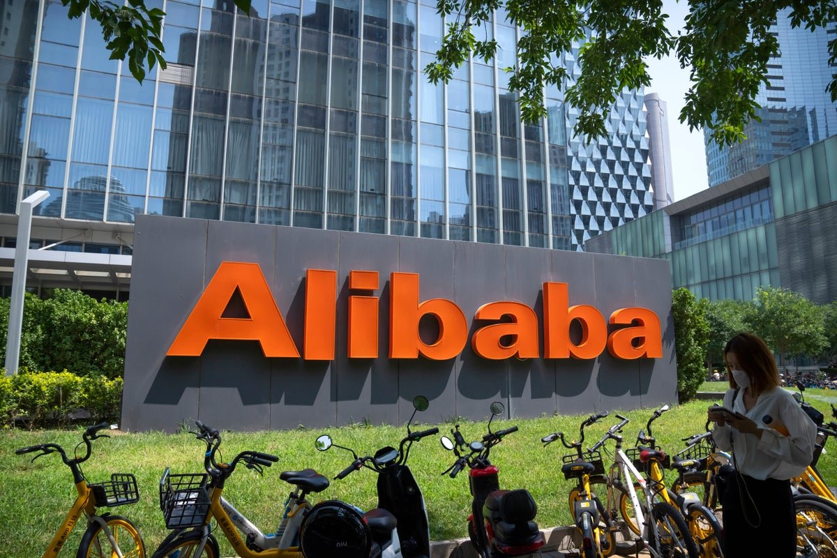 A large orange Alibaba corporate logo sign outside a modern office building with bicycles parked in front.