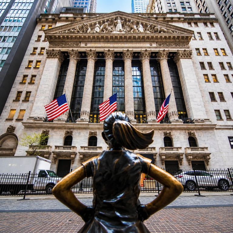 The facade of the New York Stock Exchange building adorned with American flags, viewed from behind the bronze Fearless Girl statue.