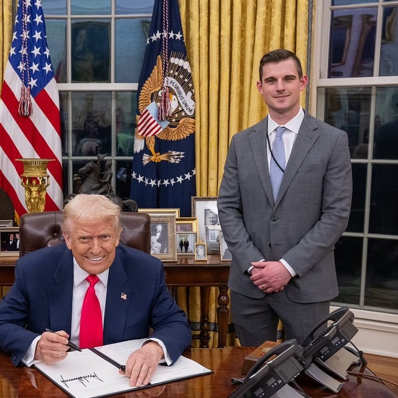 Bo Hines stands beside President Donald Trump as Trump signs documents at the Resolute Desk in the Oval Office.