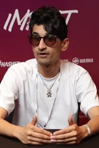 FrankDeGods sitting at a table with hands clasped, wearing sunglasses and a white T-shirt.