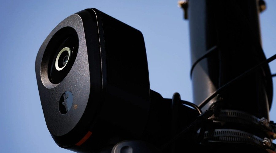 A close-up view of a Flock Safety surveillance camera mounted on a pole against a blue sky.